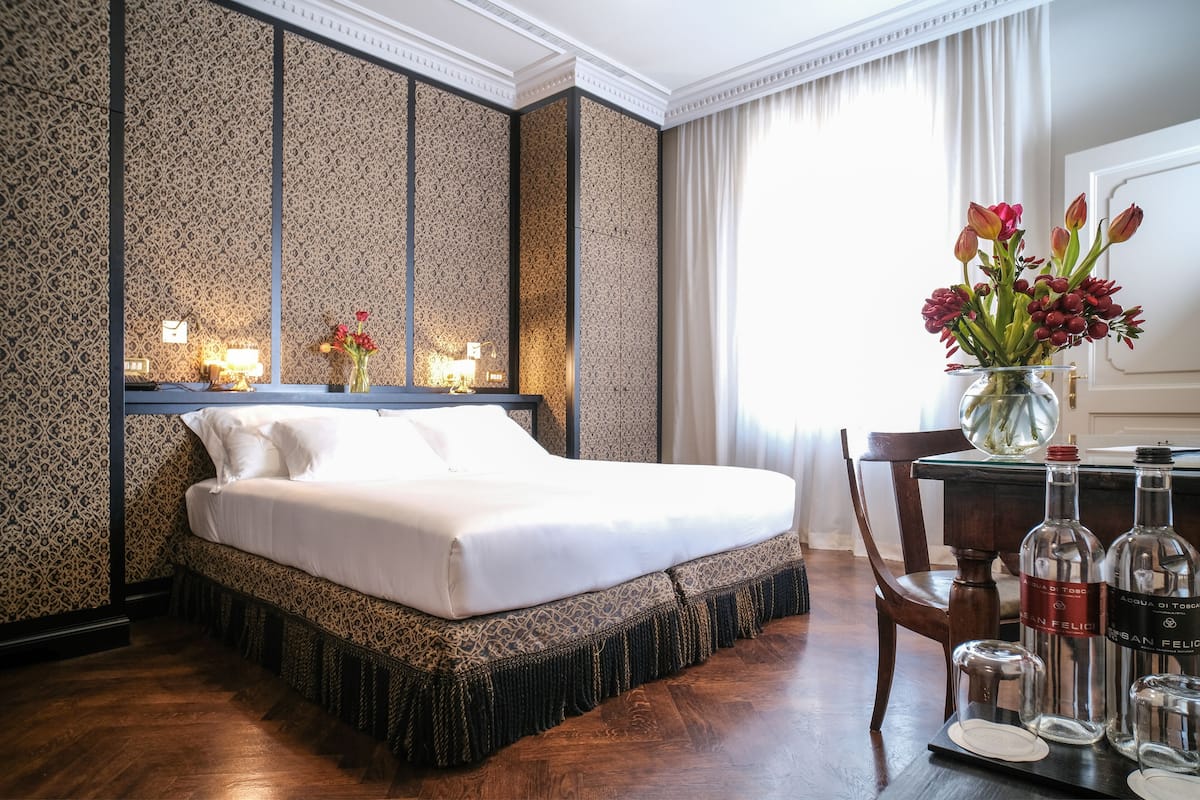 A neatly made bed in a room with patterned wallpaper, a vase of flowers, and a table with bottles and glasses.