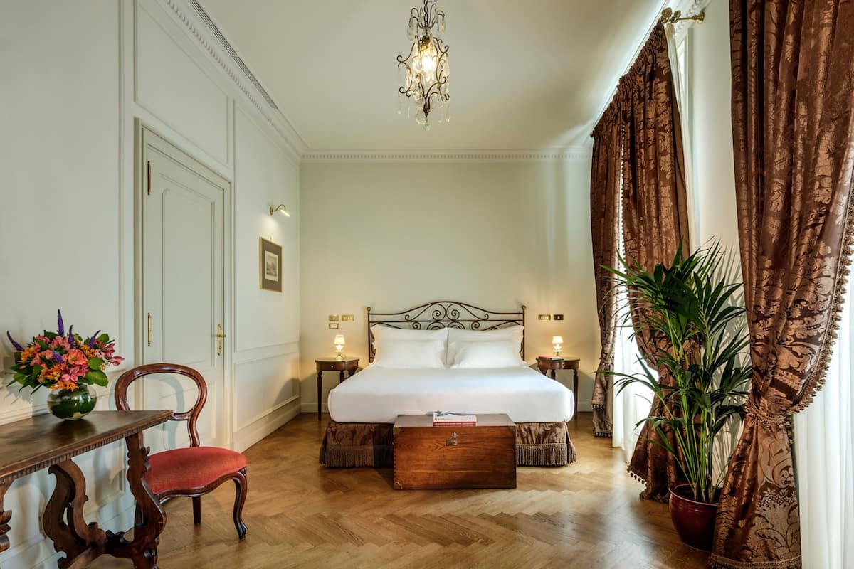 A hotel room with a large bed, a wooden chair, a bedside table, a nightstand, a chandelier, and a potted plant.