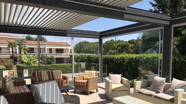 A covered outdoor seating area with wicker furniture and glass walls offering views of a residential area. - Hotel Villa Rosa (Sirmione)