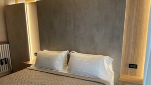 A modern bedroom with a large bed, a headboard with built-in lighting, and a radiator on the left. - Hotel Villa Rosa (Sirmione)