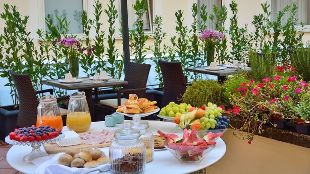 A table set with breakfast items, including fruits, pastries, and beverages, surrounded by potted plants and outdoor seating. - LH Hotel Lloyd Rome (Rome)