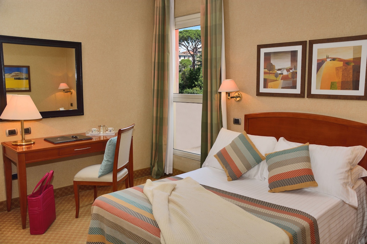 Habitación de hotel con cama, escritorio, silla, lámpara, espejo y dos cuadros en la pared.