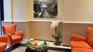 A modern lobby with orange armchairs, a glass coffee table with decorative items, a large abstract painting, and a potted plant. - LH Hotel Lloyd Rome (Rome)