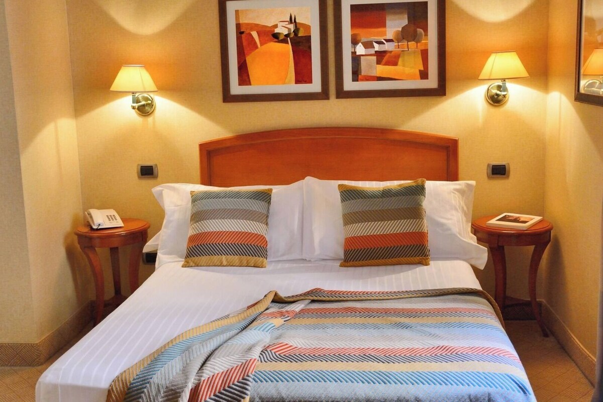 A hotel room with a bed, two bedside lamps, a telephone, and two framed artworks on the wall.
