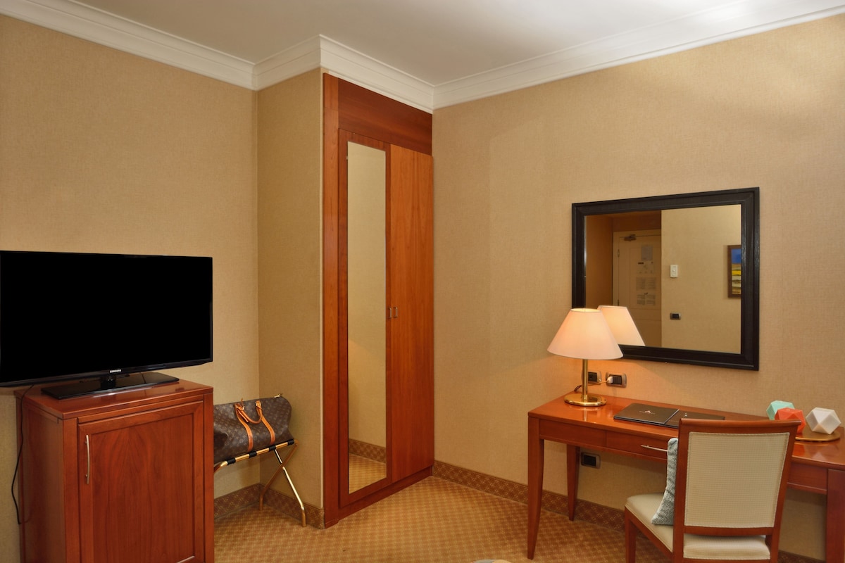 Habitación de hotel con una televisión de pantalla plana, un escritorio de madera con lámpara, una silla y un espejo en la pared.