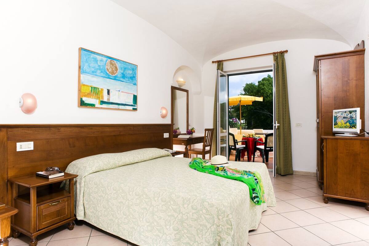 A hotel room with a bed, a nightstand, a desk, and a chair. There is a painting on the wall and a window with curtains.