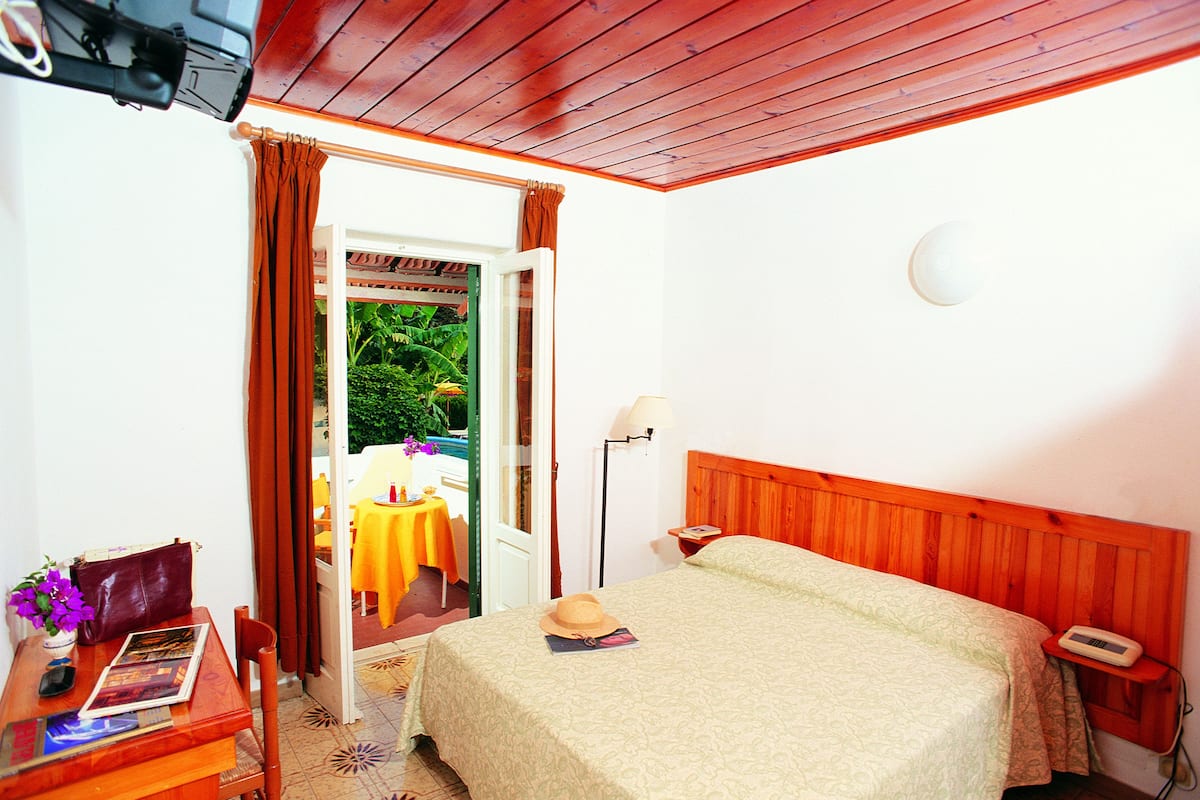 A hotel room with a bed, a bedside table, a lamp, and a view of an outdoor area with a table and chairs.