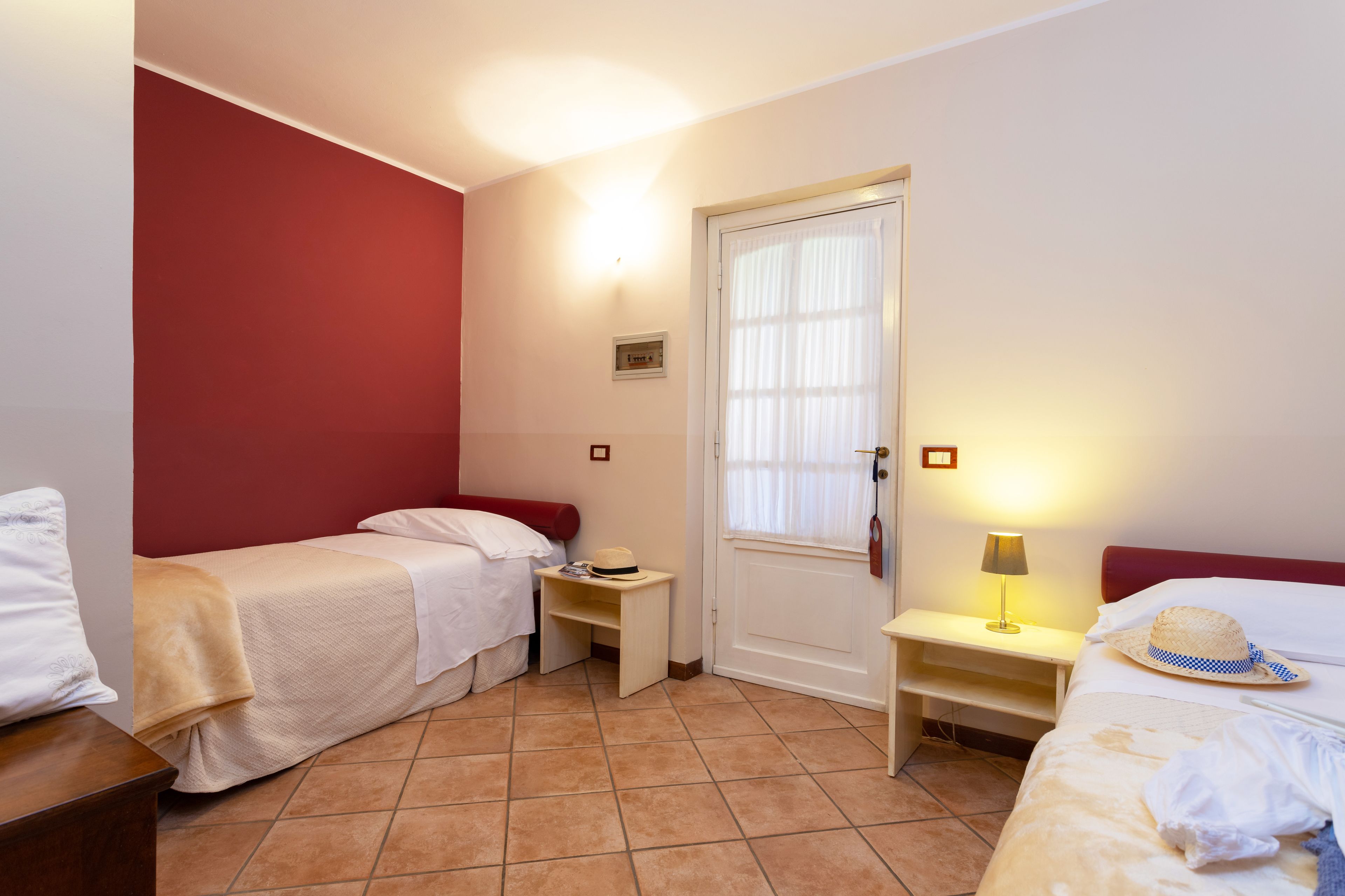 A hotel room with two single beds, a small bedside table with a lamp, a door with a window, and a red accent wall.