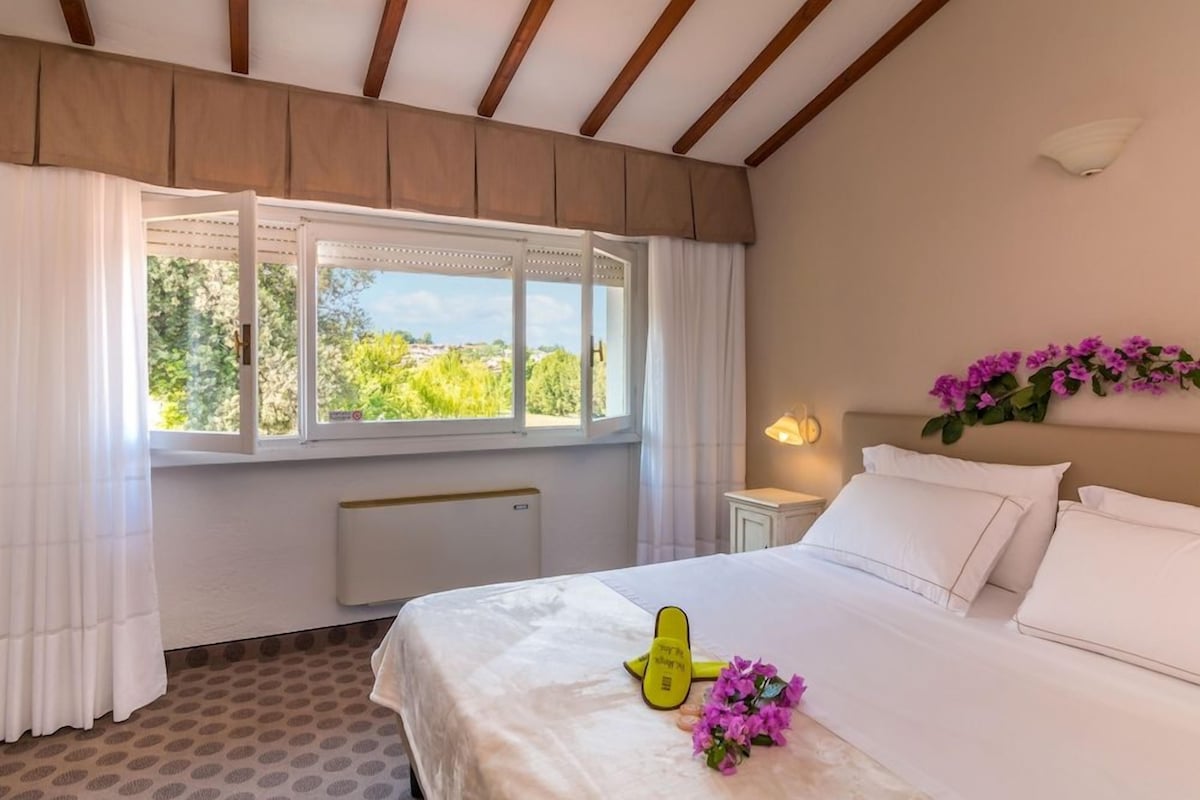 Un dormitorio con cama, ventana con cortinas, mesita de noche con lámpara y vistas al exterior con vegetación.