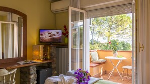 Habitación de hotel con cama, televisor, una mesita con un jarrón de flores, una silla y balcón con vistas a árboles.