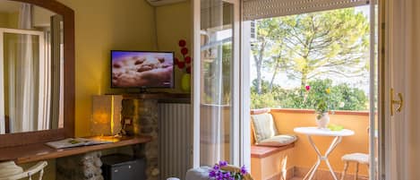 A hotel room with a bed, a TV, a small table with a vase of flowers, a chair, and a balcony with a view of trees.