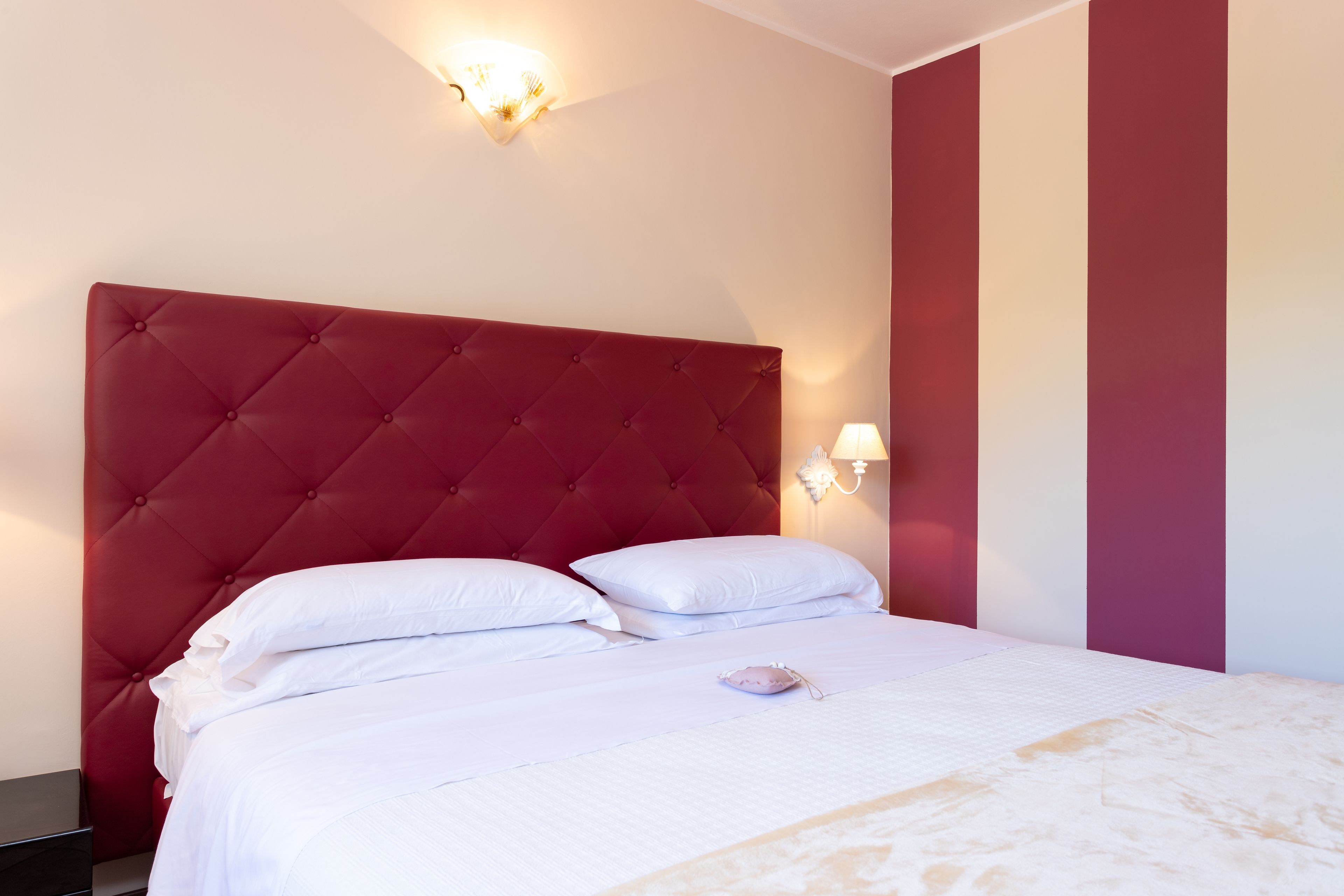 Un lit bien fait, avec du linge de lit blanc et une tête de lit rouge, une lampe de chevet et un mur orné de rayures verticales.