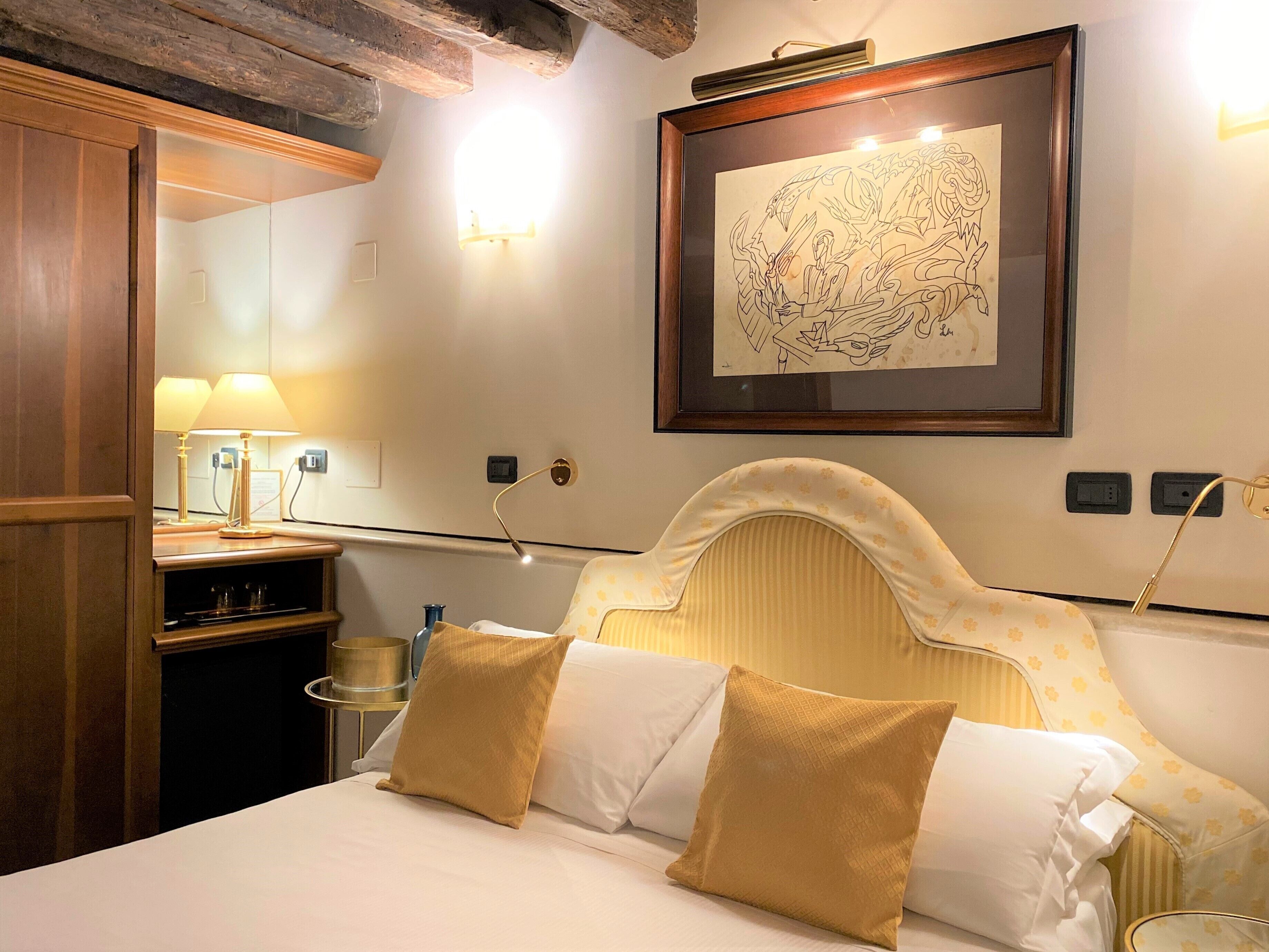 A hotel room with a bed, a framed artwork on the wall, a bedside lamp, and a wooden headboard.