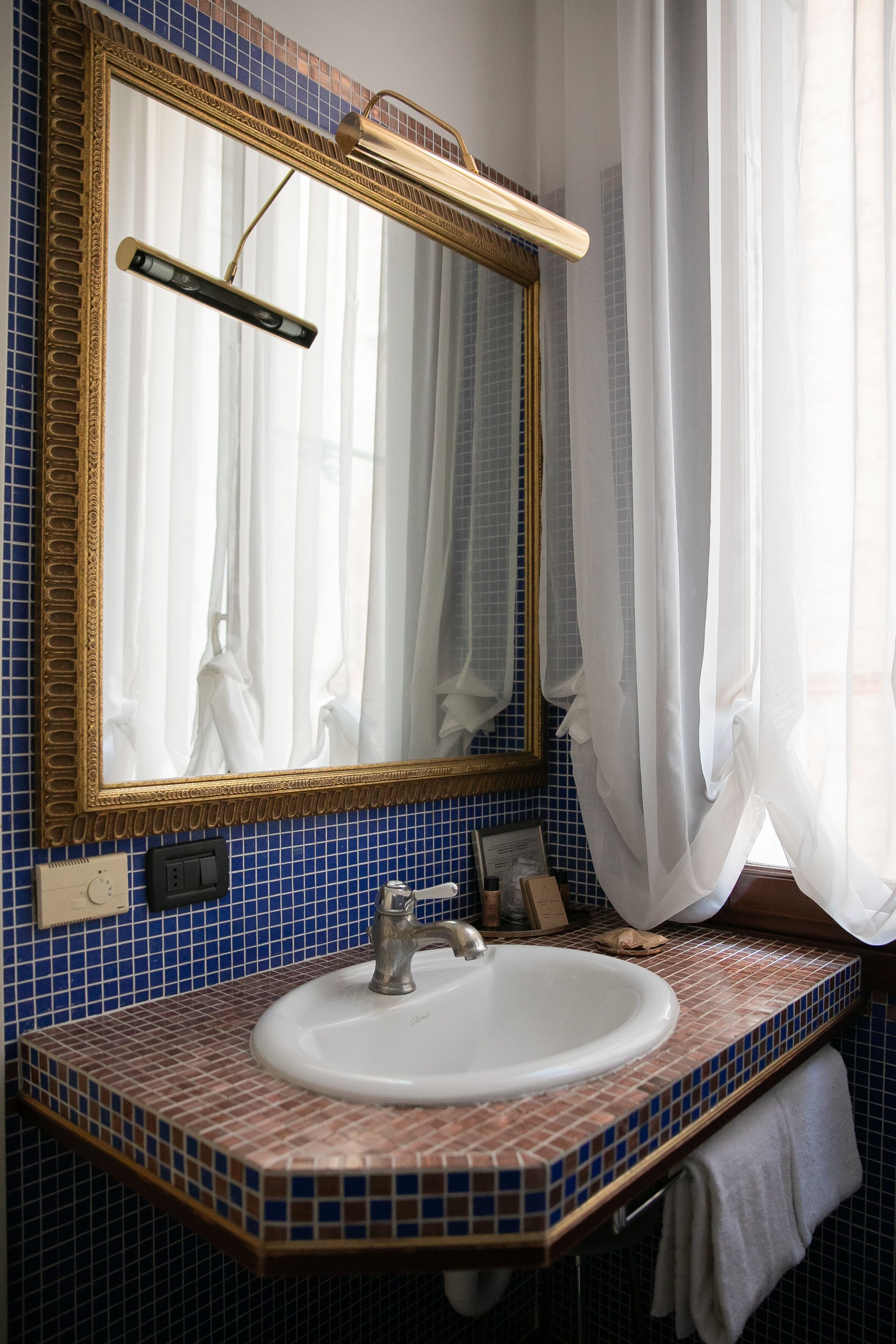 Une salle de bain avec un mur recouvert de carreaux bleus, un lavabo blanc, un miroir à cadre doré et une serviette accrochée au mur.