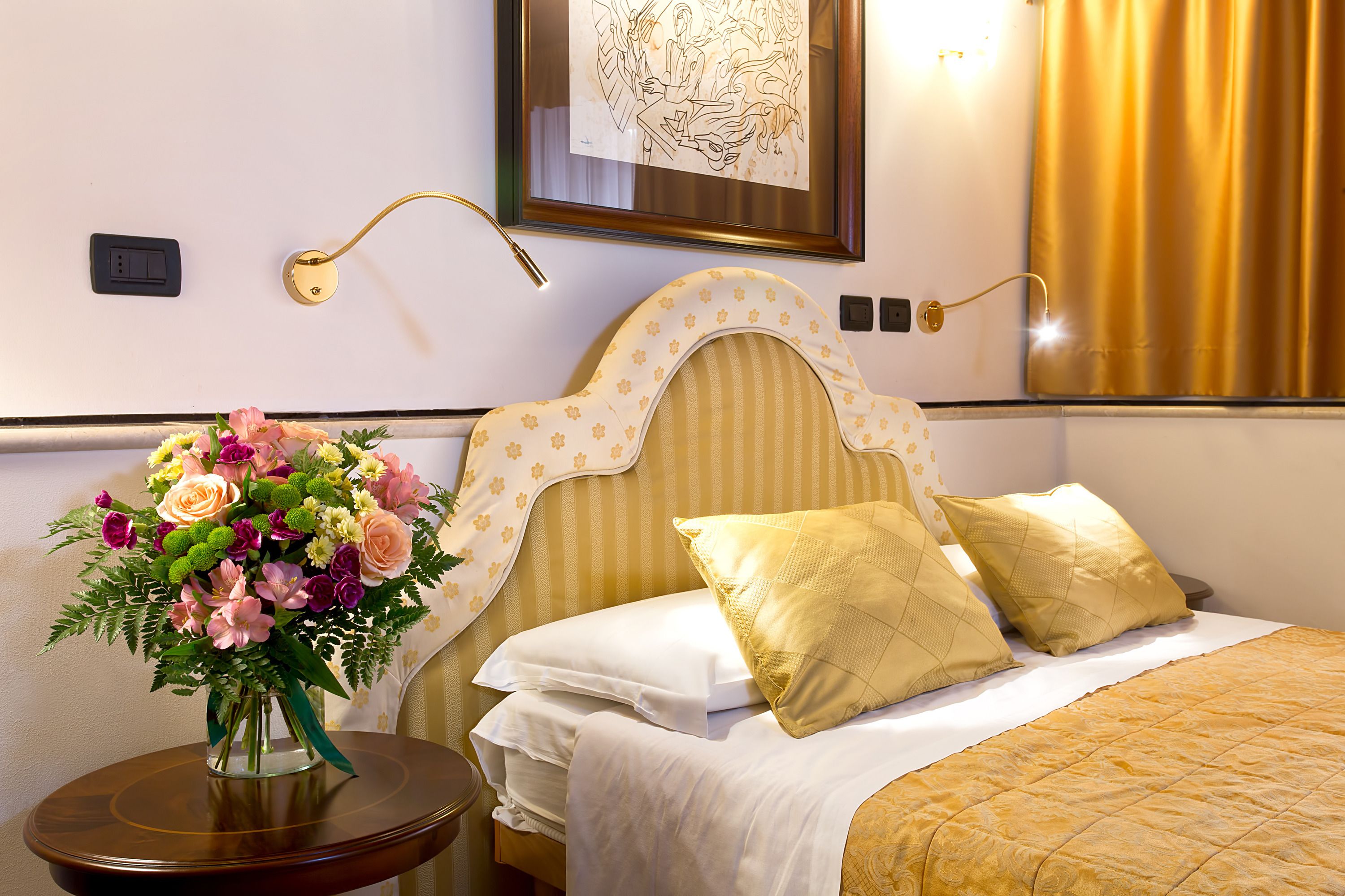 A hotel room with a bed, a nightstand with a vase of flowers, and a wall-mounted lamp.