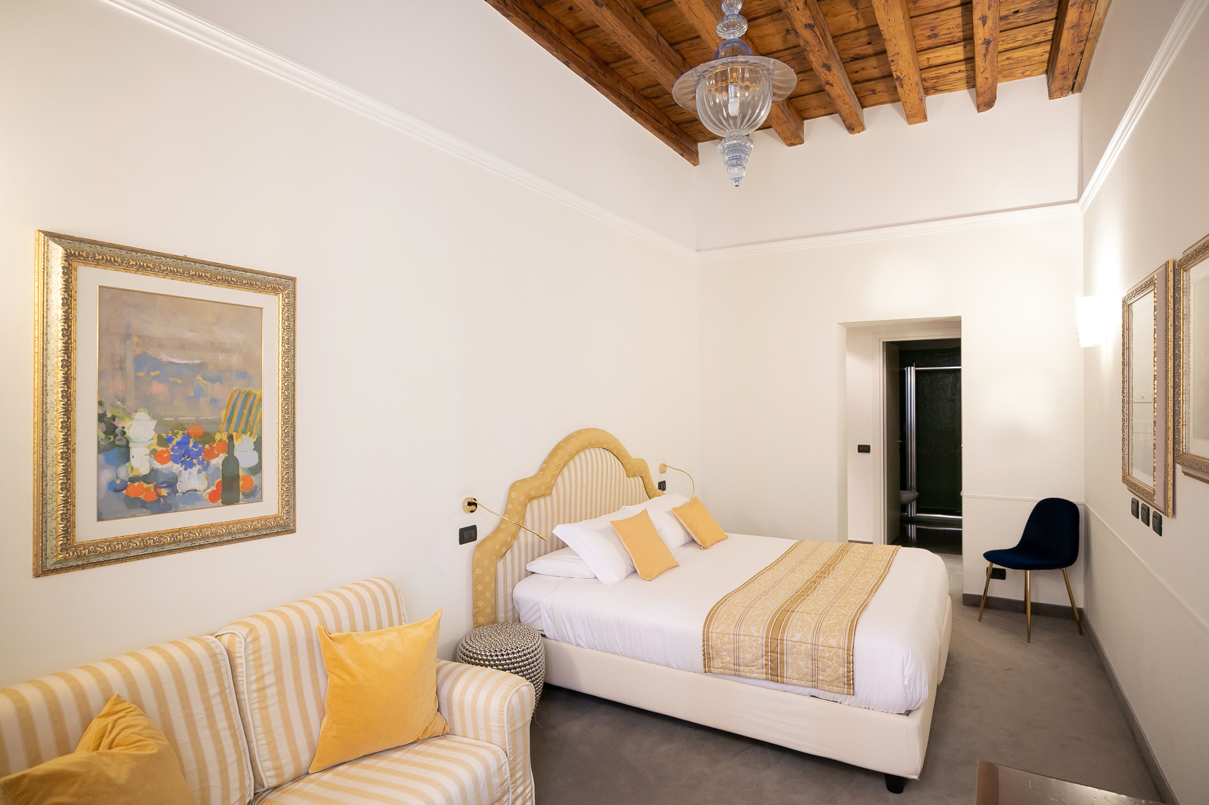 A bedroom with a bed, a sofa, a chair, a framed painting, and a chandelier.