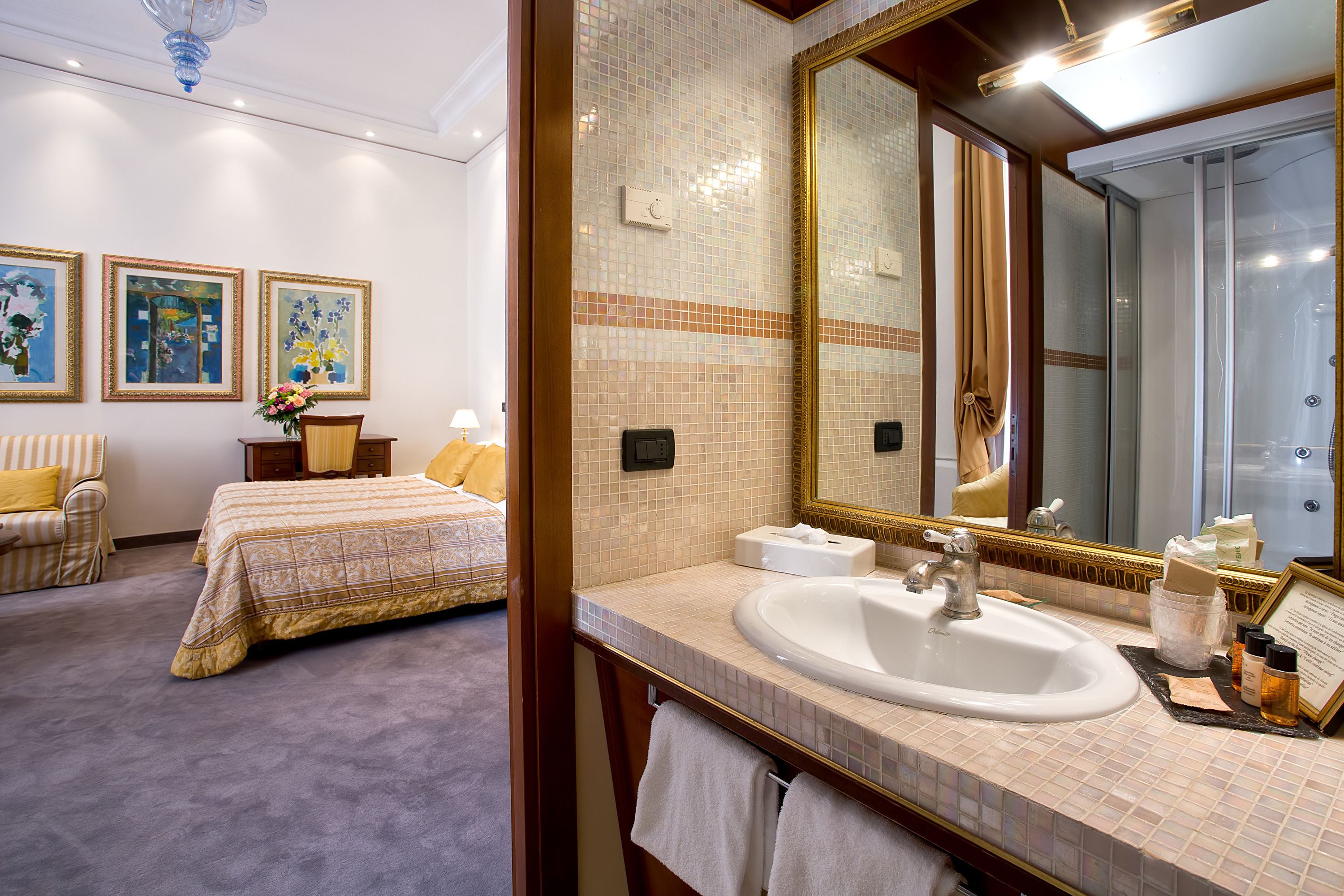 Una habitación de hotel con una cama grande, un espacio de descanso con una silla y una mesa, un baño con lavamanos y espejo, y obras de arte en las paredes.