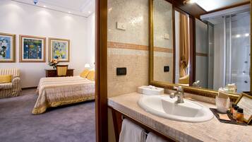 Een hotelkamer met een groot bed, een zithoek met een stoel en tafel, een badkamer met wastafel en spiegel, en kunstwerken aan de muren.