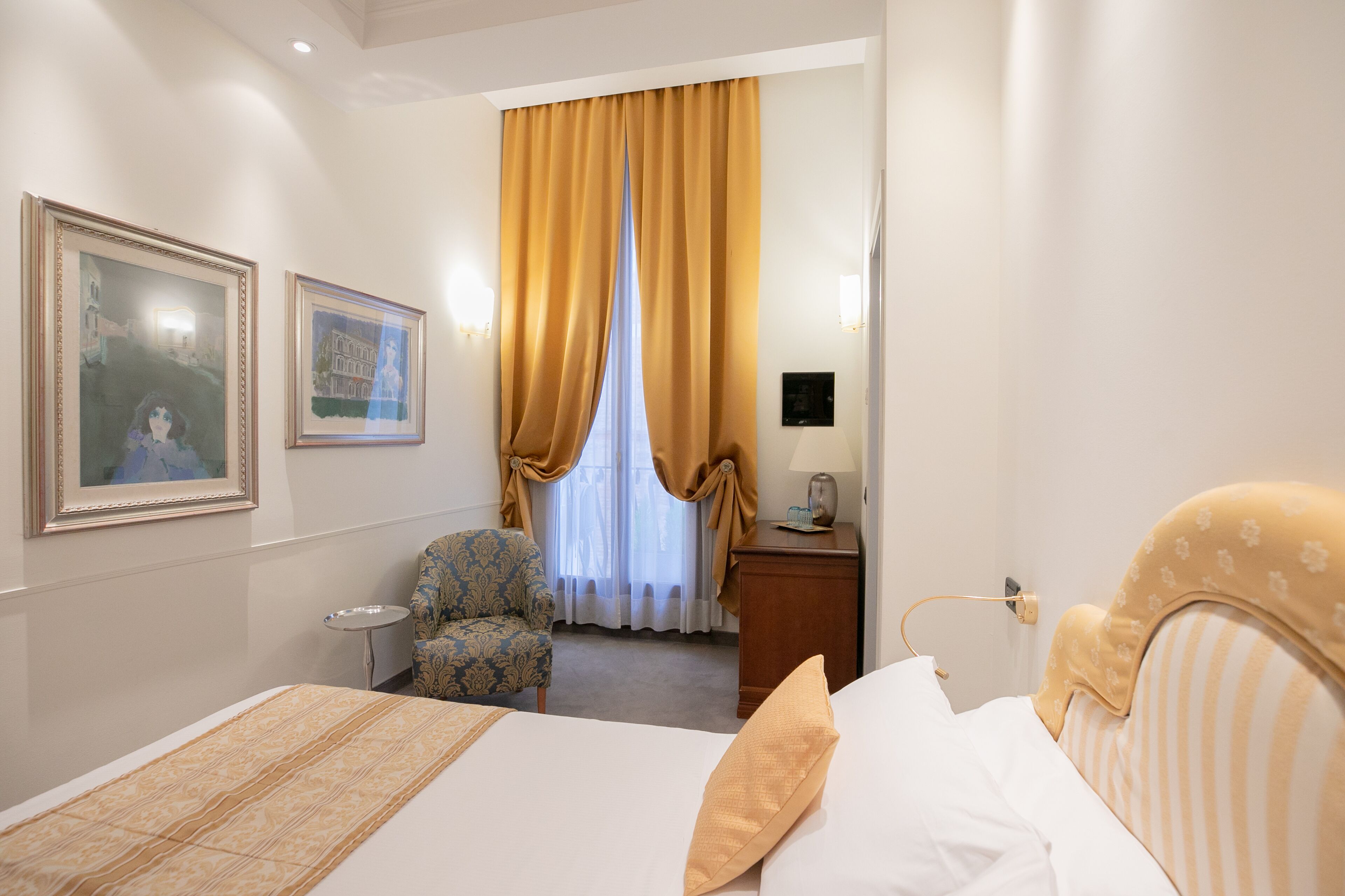 A hotel room with a bed, a chair, a desk, and two framed pictures on the wall.