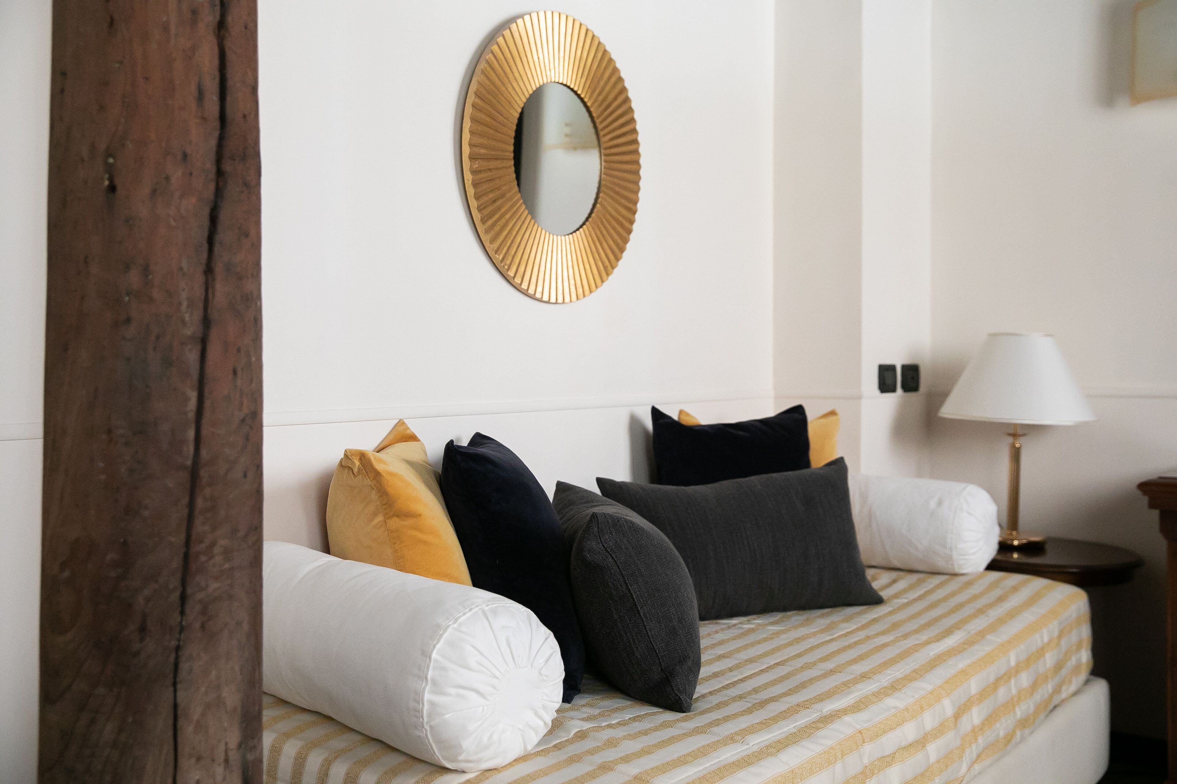 A neatly made bed with striped bedding, a round mirror with a gold frame, and a bedside lamp.