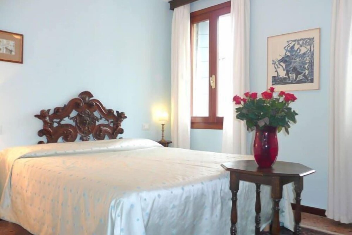 Une chambre d’hôtel avec un lit, une tête de lit en bois, une fenêtre avec des rideaux, un vase avec des fleurs et un tableau encadré au mur.