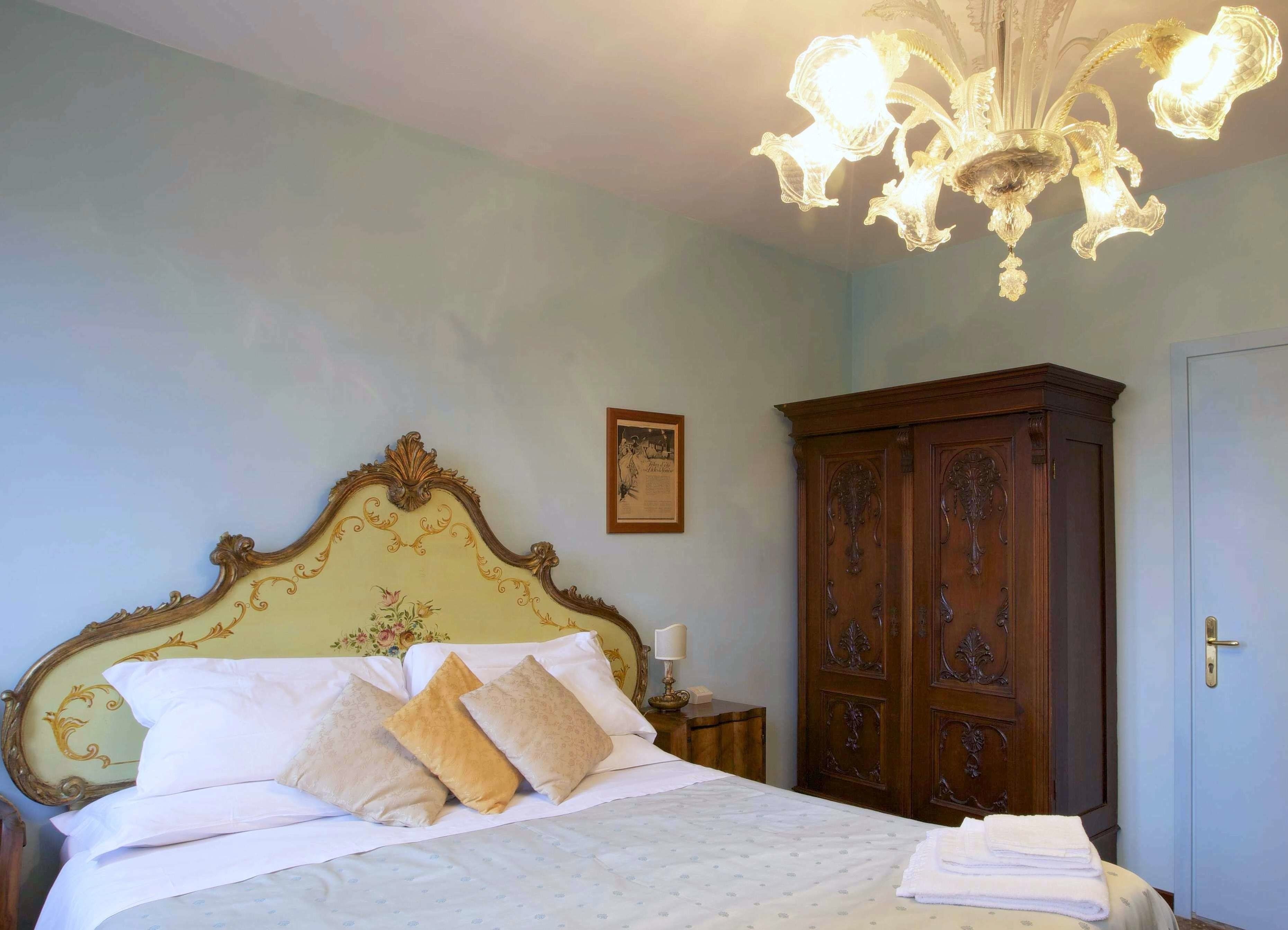 A bedroom with a large bed, ornate headboard, a chandelier, a wooden wardrobe, and a framed certificate on the wall.