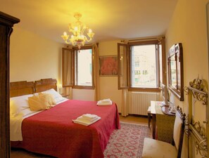 A hotel room with a large bed, a chandelier, a window with a view of buildings, a radiator, and a desk with a mirror. - 3749 Ponte Chiodo Guest House (Venice)