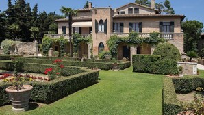 Un edificio grande e histórico con fachada simétrica, ventanas arqueadas y un jardín cuidado con setos y macizos de flores.