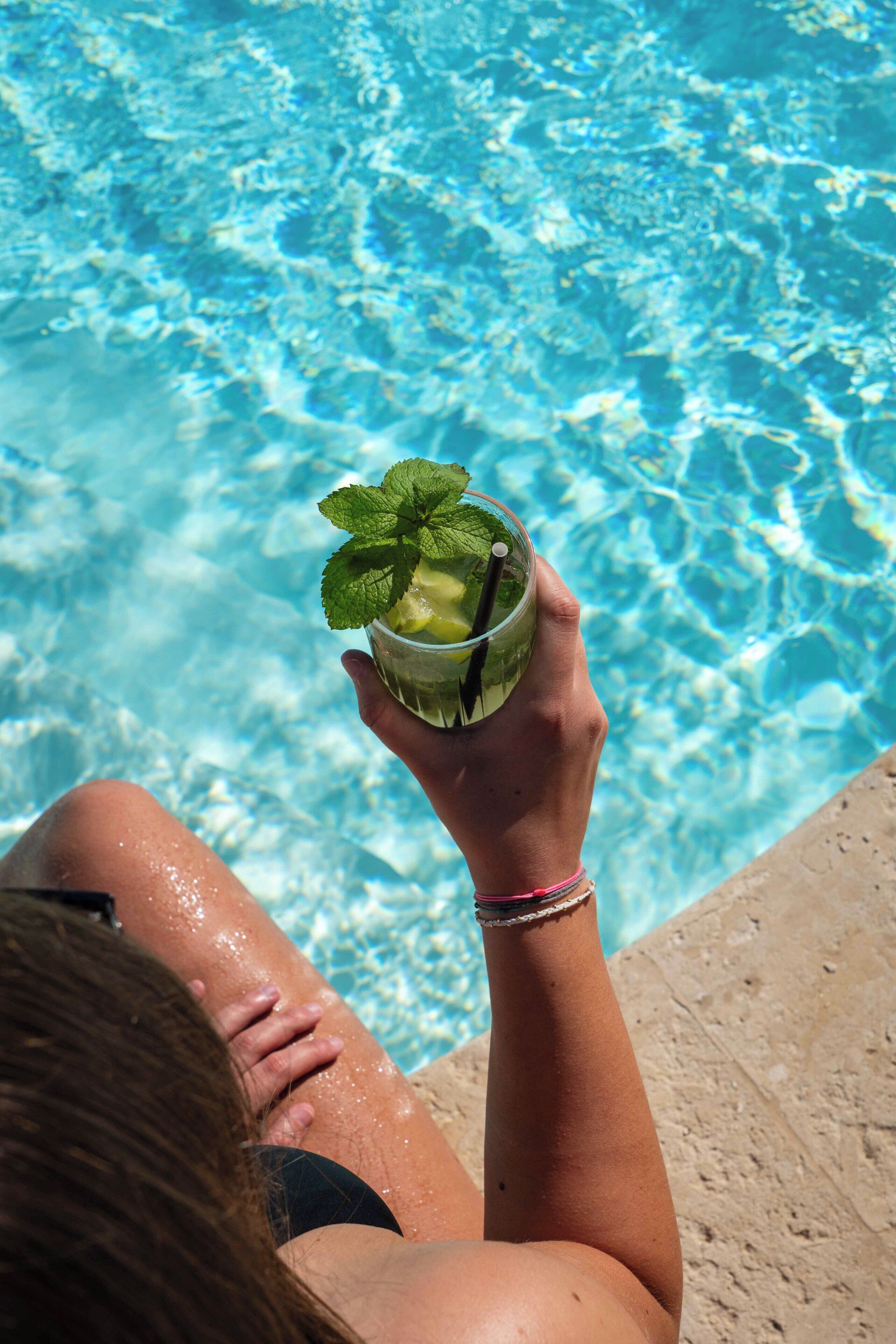 A person holding a drink with a mint garnish by a pool.