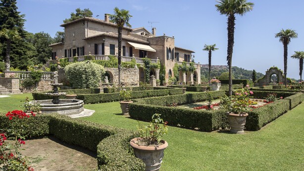 Un edificio grande e histórico con un jardín bien cuidado que incluye setos, una fuente y palmeras.