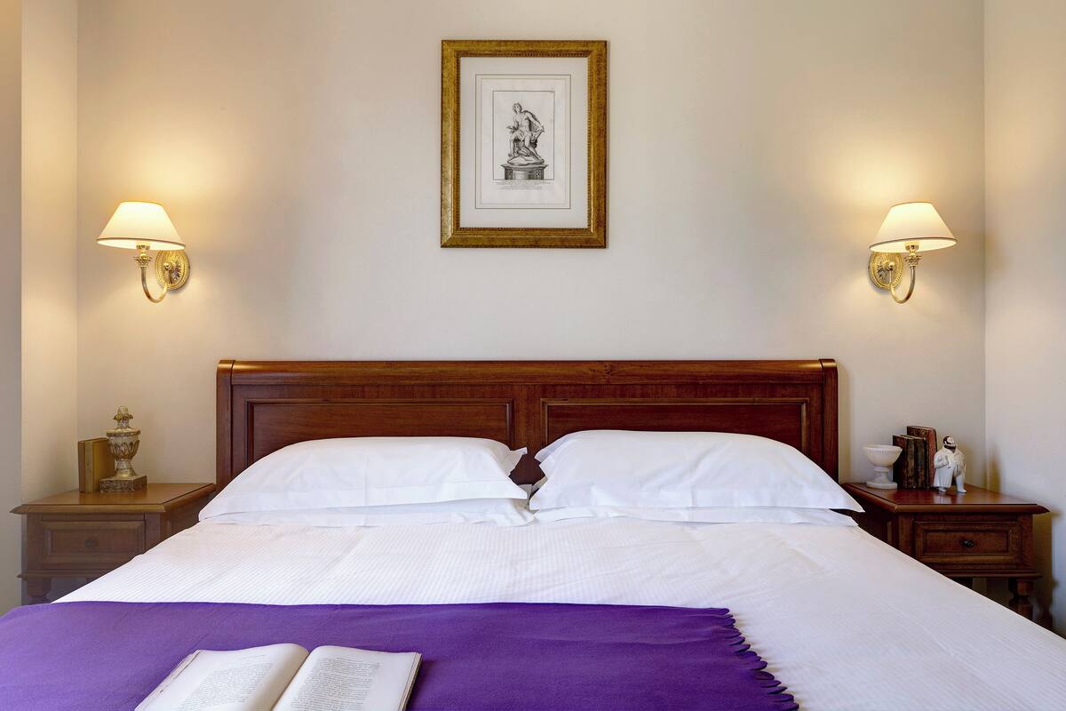 A neatly made bed with white linens and a purple throw, flanked by two bedside tables with lamps and a framed artwork above.