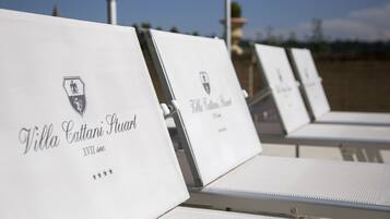 Filas de sillas blancas de descanso con la marca 'Villa Cattani Stuar' y un escudo, dispuestas al aire libre con cielo despejado al fondo.