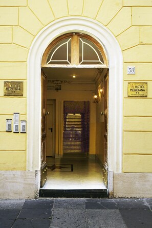 La entrada del Hotel Meridiana, con un arco clásico, un piano en el segundo piso y un cartel que indica el nombre y la ubicación del hotel.