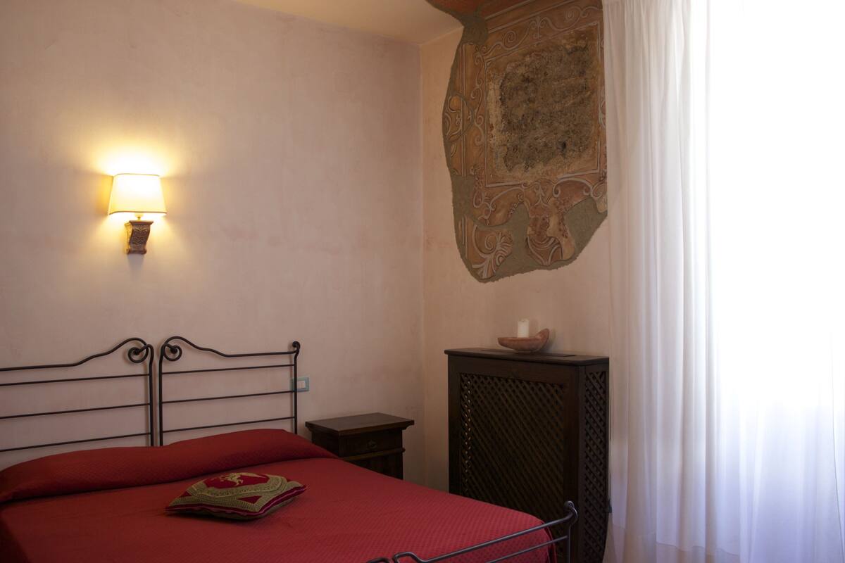 A bedroom with a metal bed frame, a red bedspread, a wall-mounted lamp, a small wooden nightstand, and a decorative wall piece.