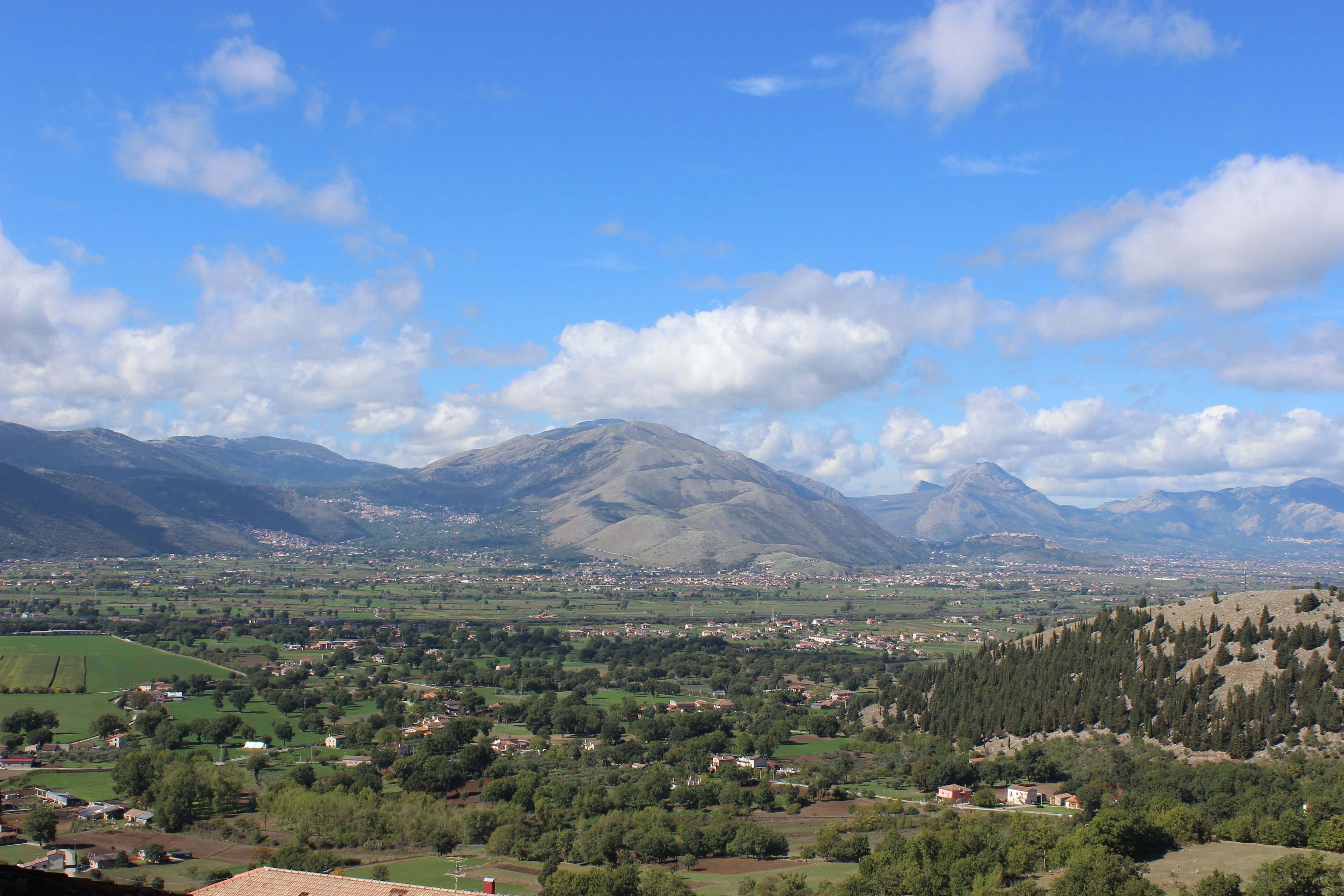 A panoramic view of a valley with mountains in the background, a town in the middle, and a mix of green fields and buildings.
