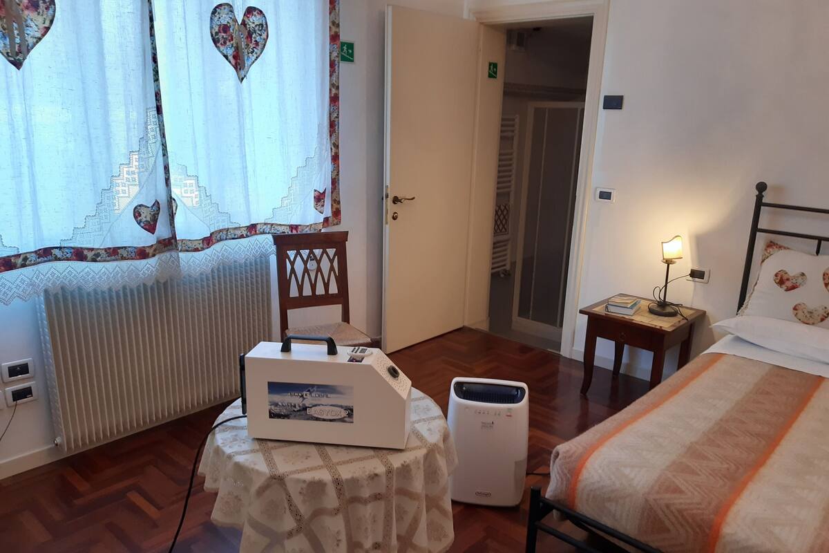 Habitación de hotel con una cama, una mesa con una computadora portátil, una silla, un radiador, una mesita de noche con una lámpara y una puerta que da a un pasillo.