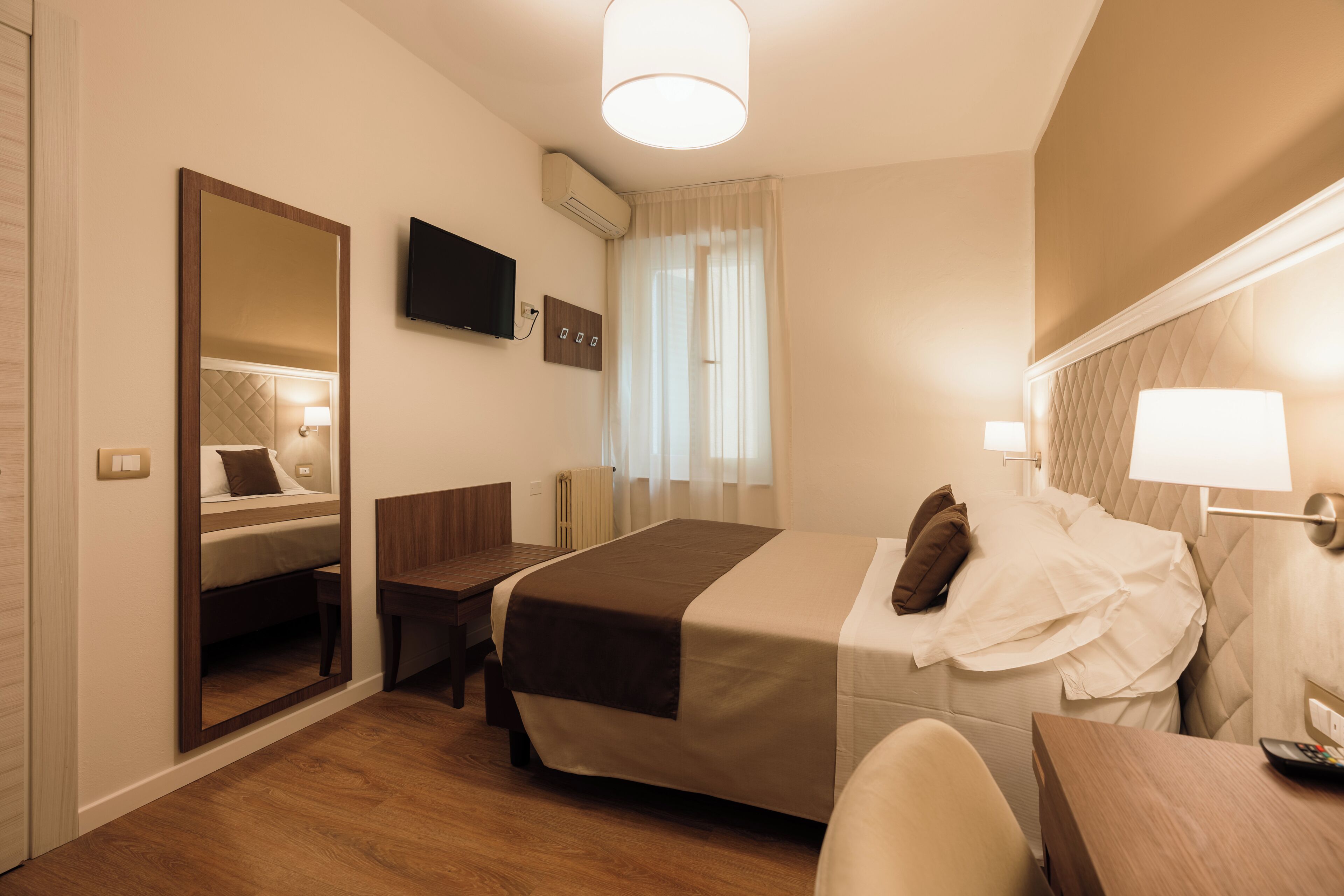 Quarto de hotel com cama, mesa de cabeceira, cadeira, escrivaninha, espelho, televisão e janela com cortinas.