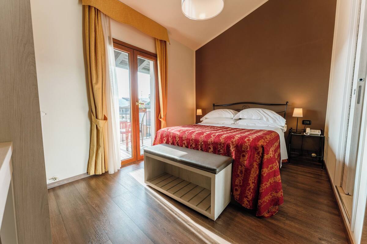 Un dormitorio con cama, mesitas de noche, un banco, una ventana con cortinas y vistas al exterior.