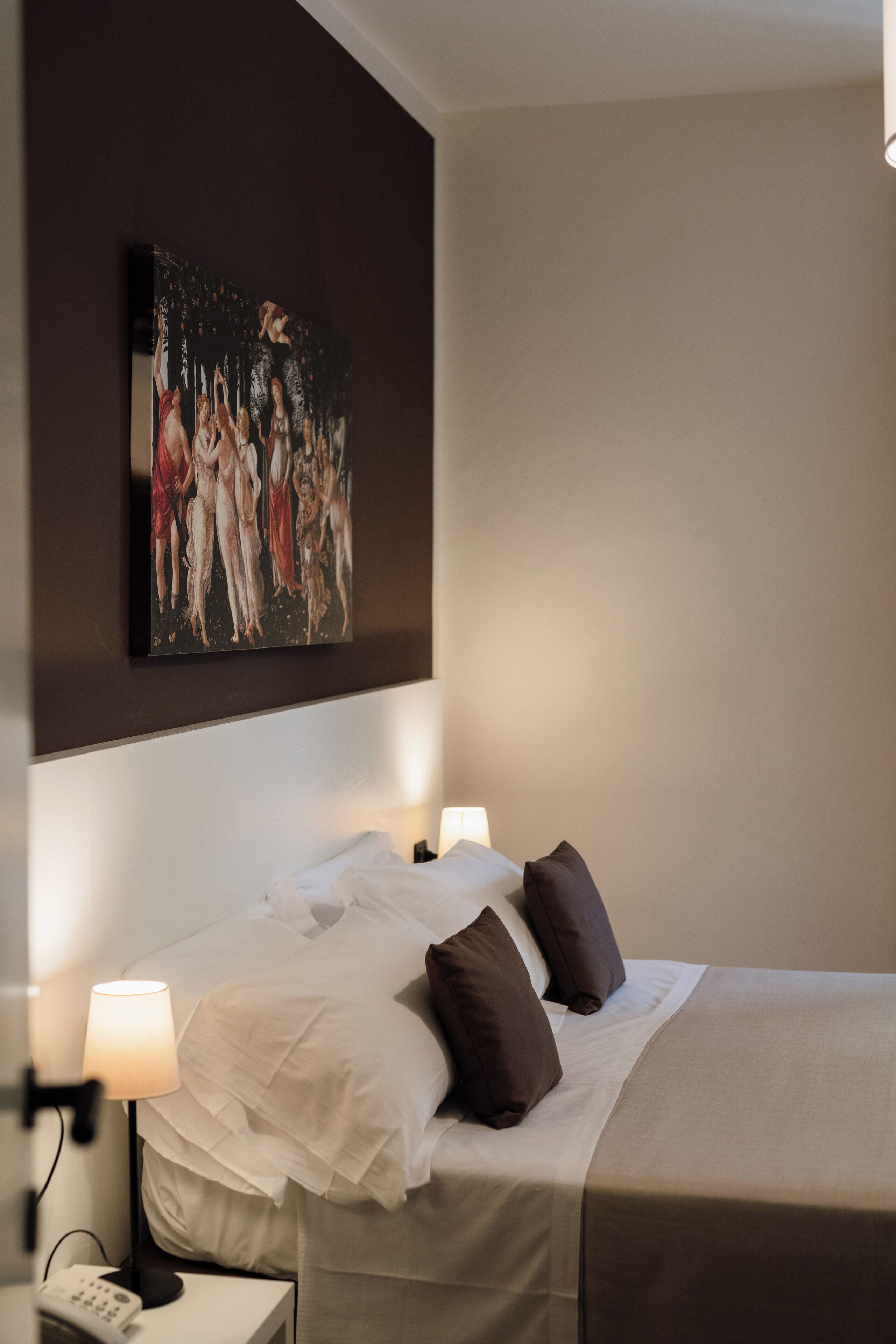 A hotel room with a bed, two bedside lamps, a painting on the wall, and a nightstand with a phone.