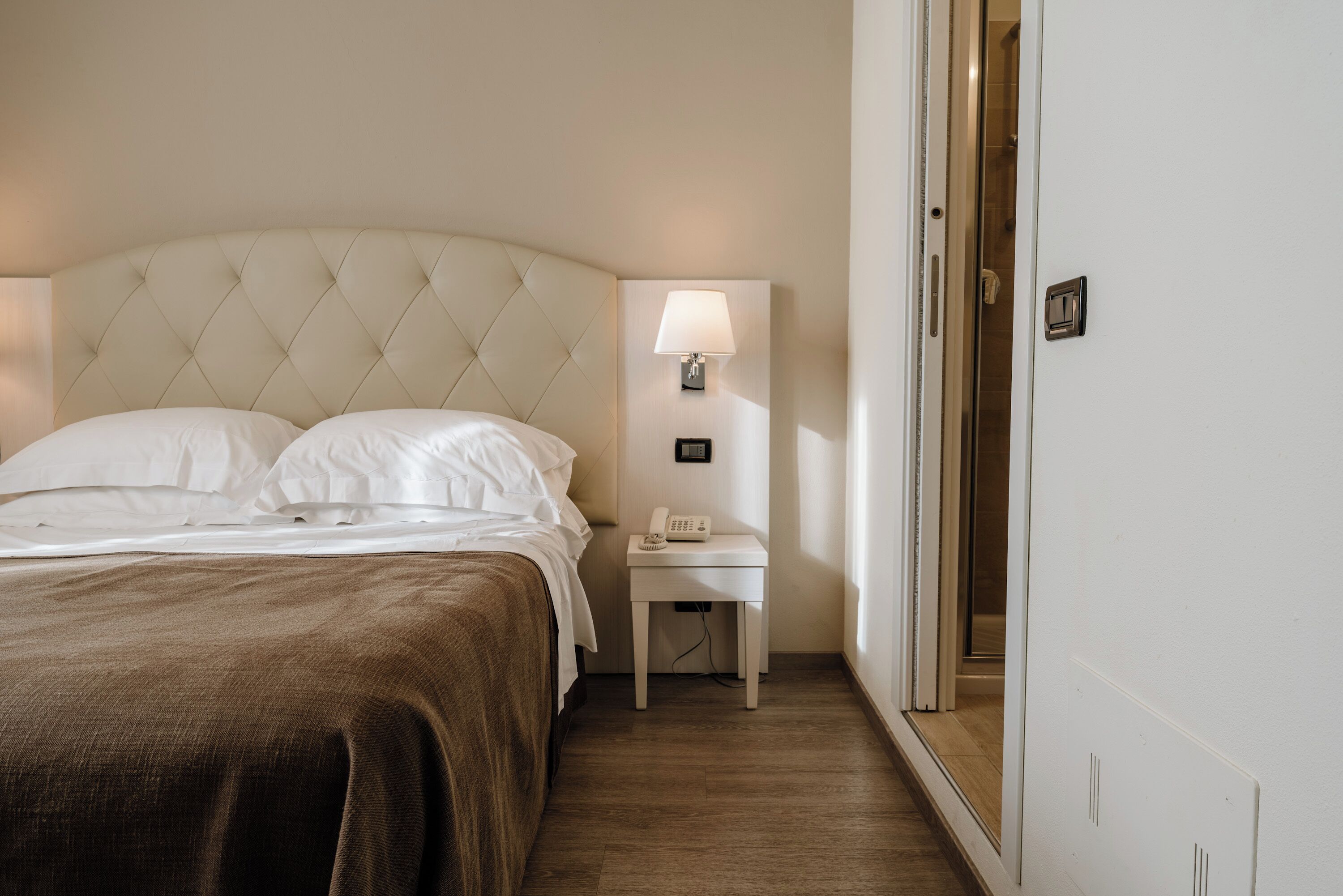Ein ordentlich bezogenes Bett mit einer braunen Tagesdecke, ein Nachttisch mit Lampe und Telefon.