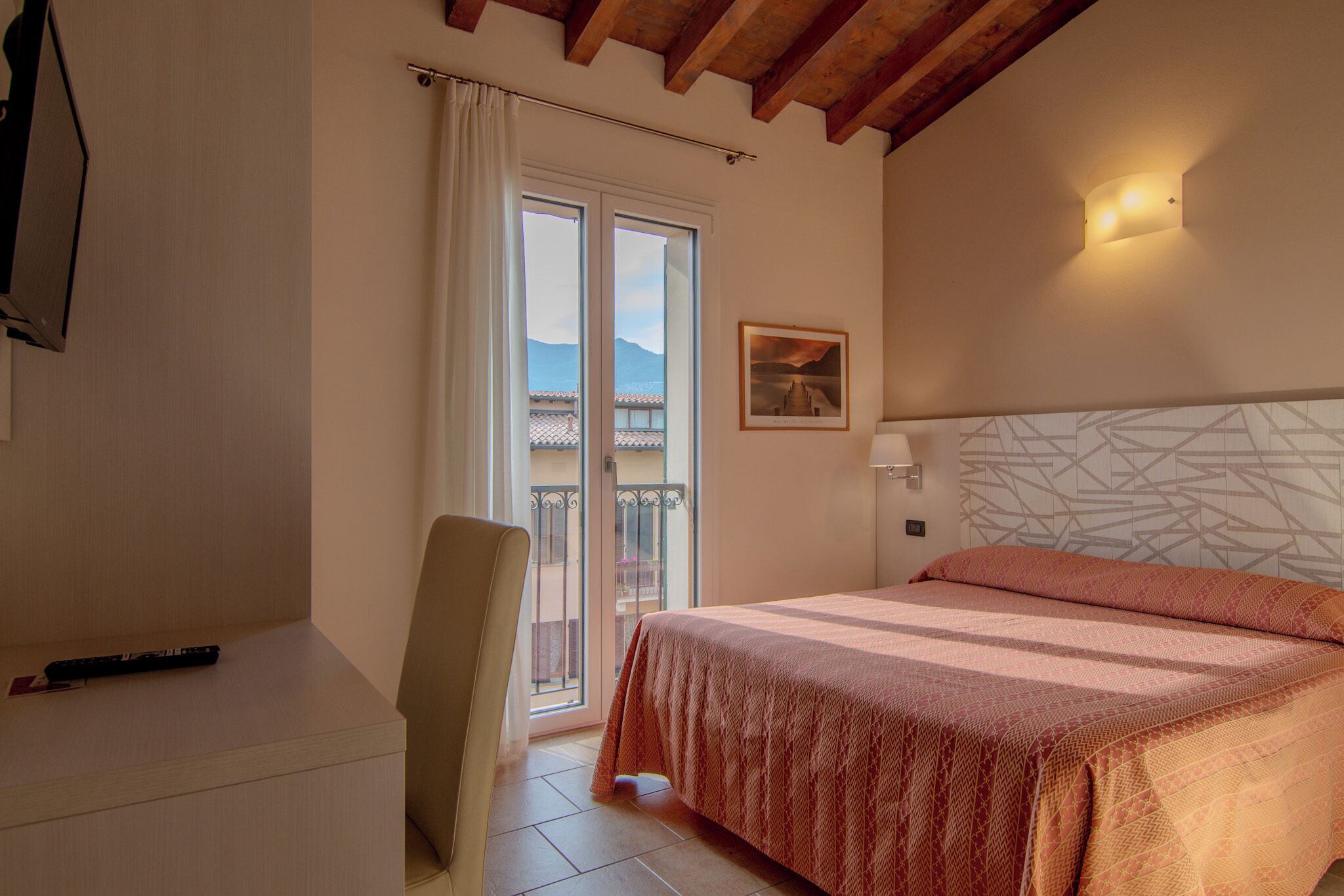 Un dormitorio con cama, una silla, un escritorio, una ventana con vistas a montañas y una lámpara fijada a la pared.
