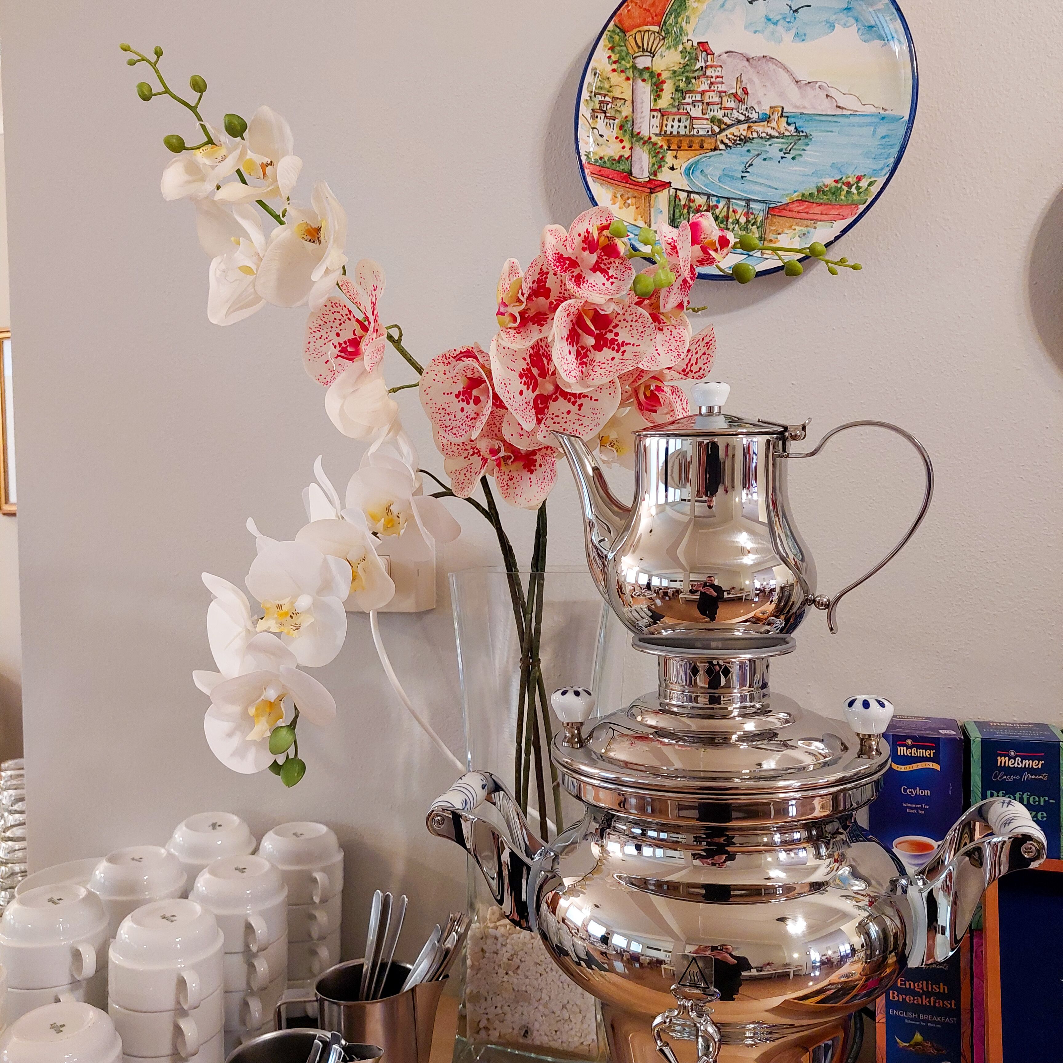 A room with a decorative orchid arrangement, a silver teapot, and a plate with a coastal scene.