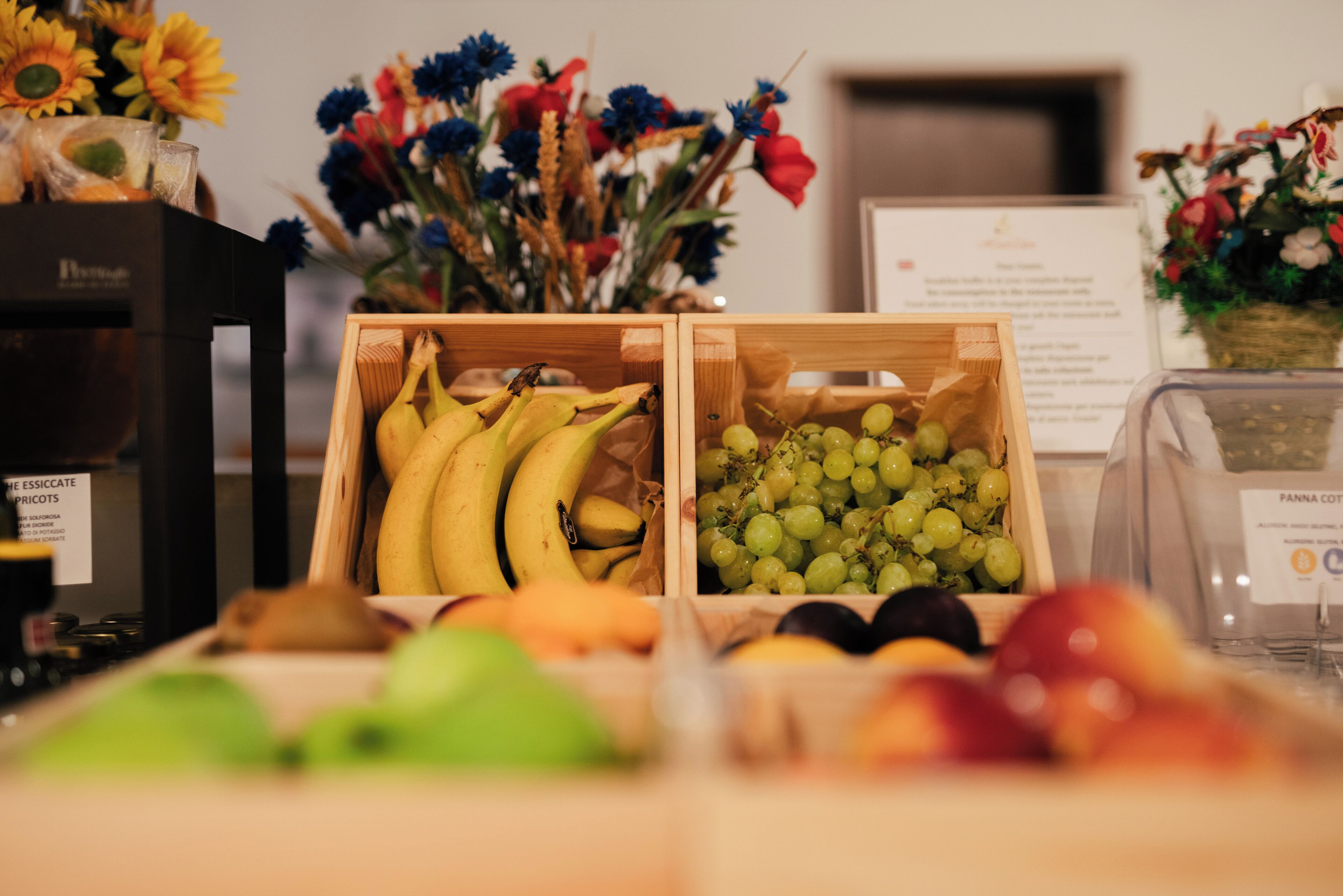 Une présentation de fruits comprenant des bananes et des raisins dans des caisses en bois, une machine à café et, au fond, un vase rempli de fleurs.