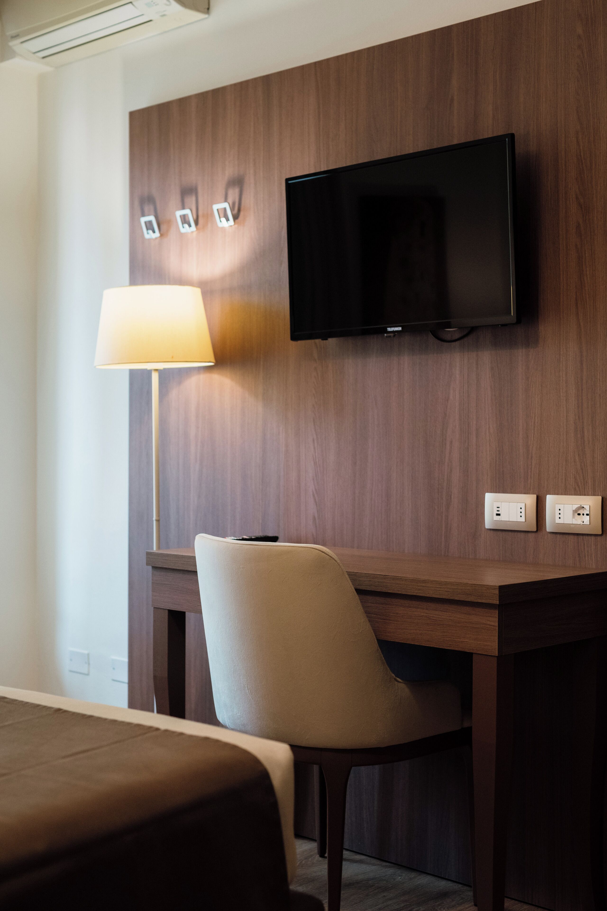 A hotel room with a bed, a desk with a chair, a television mounted on the wall, and a lamp.