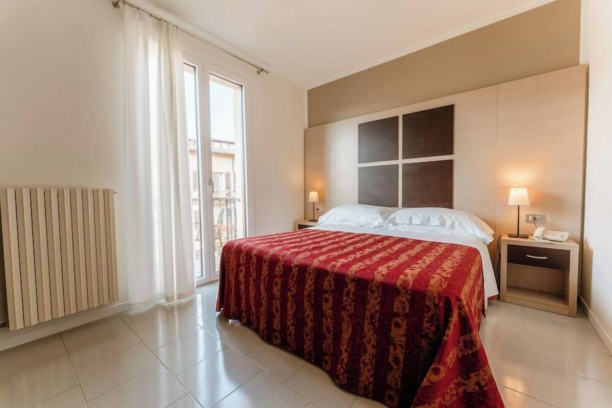 Habitación de hotel con cama, mesita de noche, lámpara, ventana con cortinas y radiador.