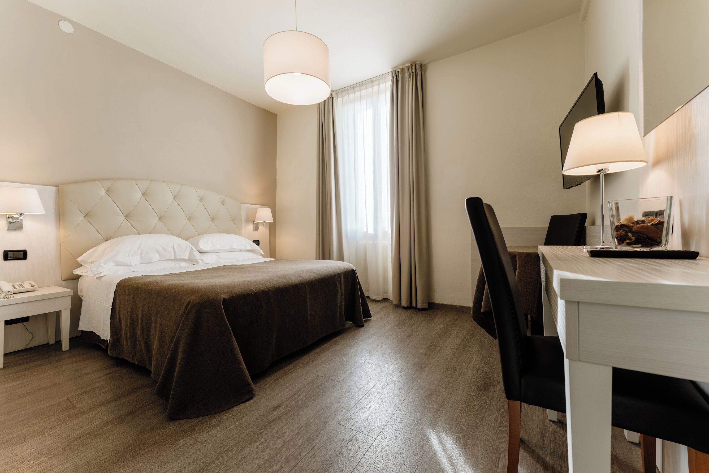 Una habitación de hotel con cama, mesita de noche, lámpara, silla, escritorio y un amplio ventanal con cortinas.