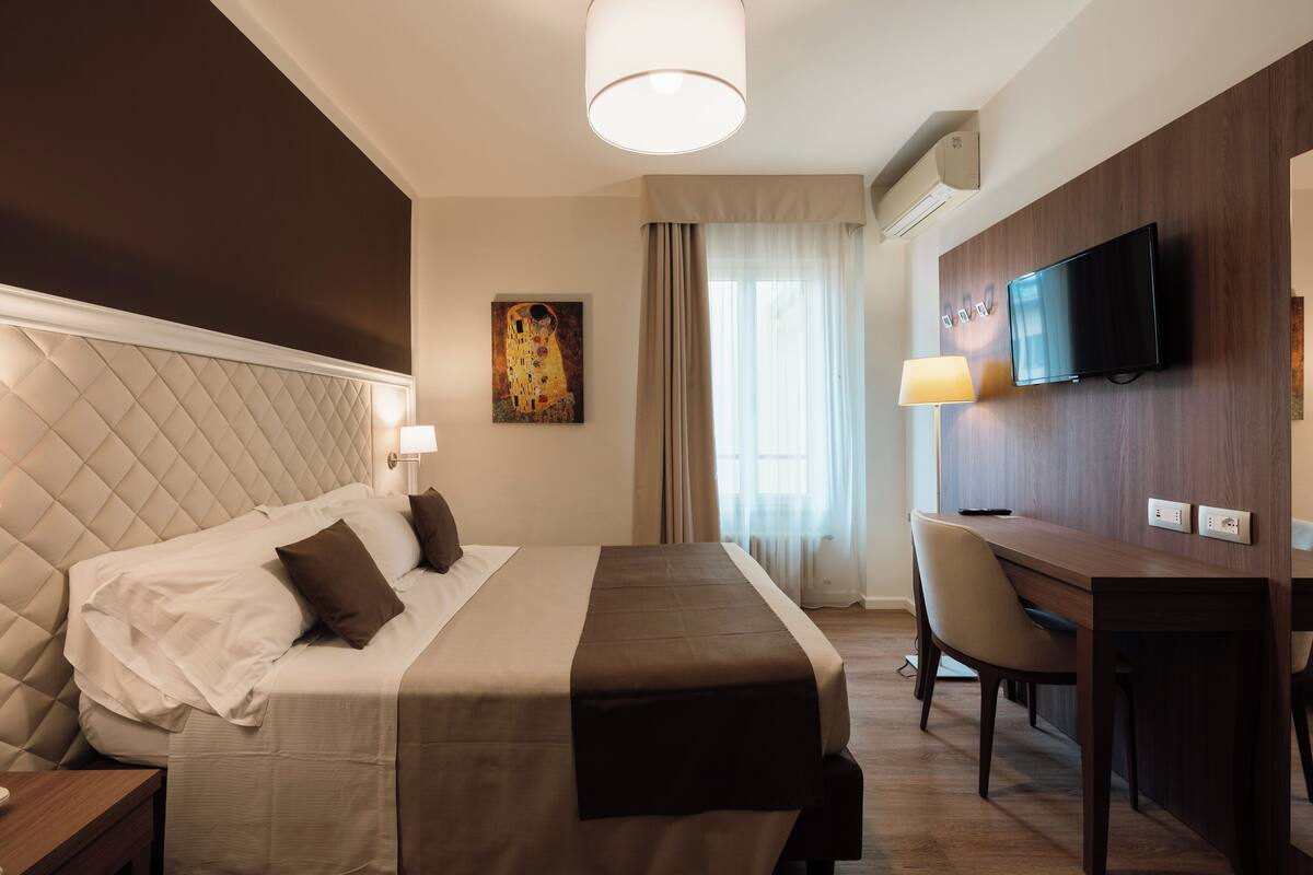 Una habitación de hotel con una cama, un escritorio, una silla, un televisor y un cuadro en la pared.