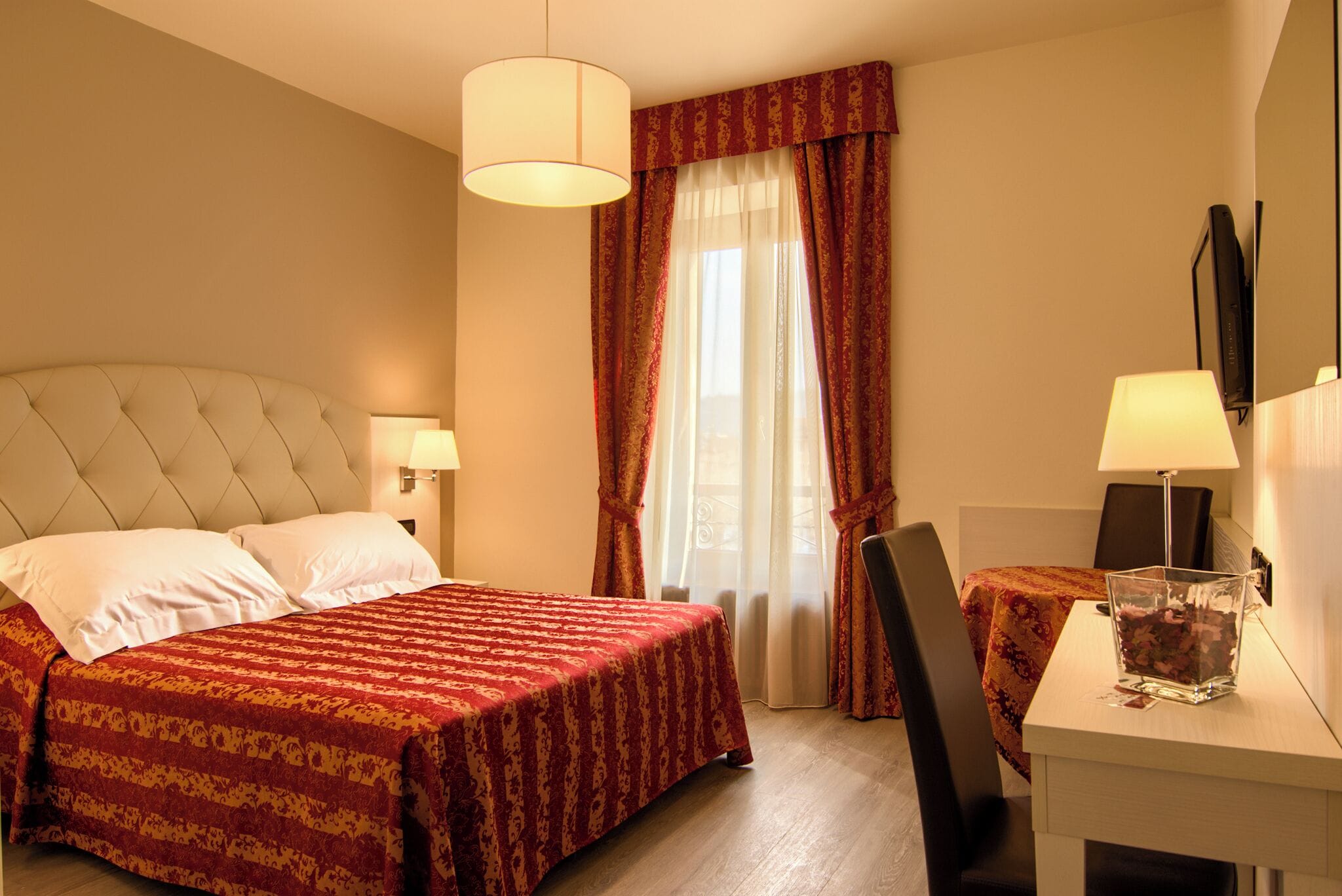 Camera d'albergo con un letto, due lampade sui comodini, una scrivania con una sedia e un'ampia finestra con tende.