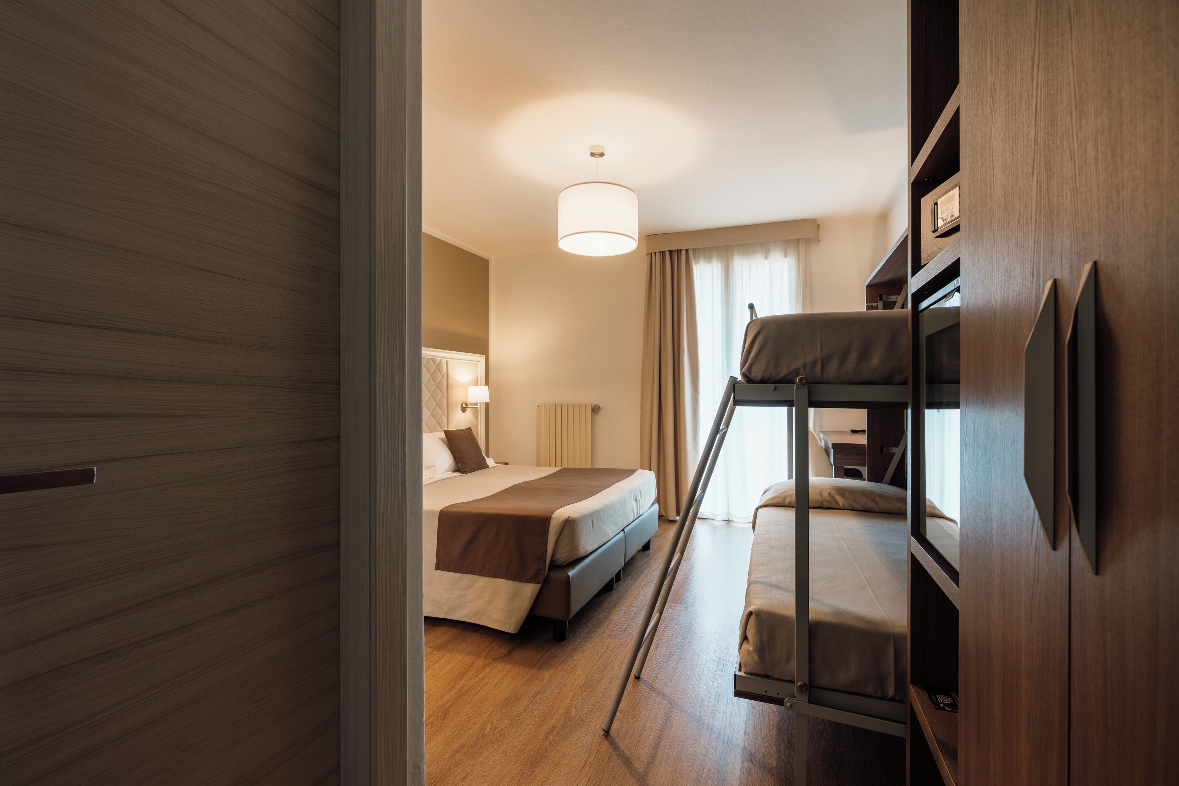 A hotel room with a bunk bed, a single bed, a bedside table with a lamp, and a wooden wardrobe.