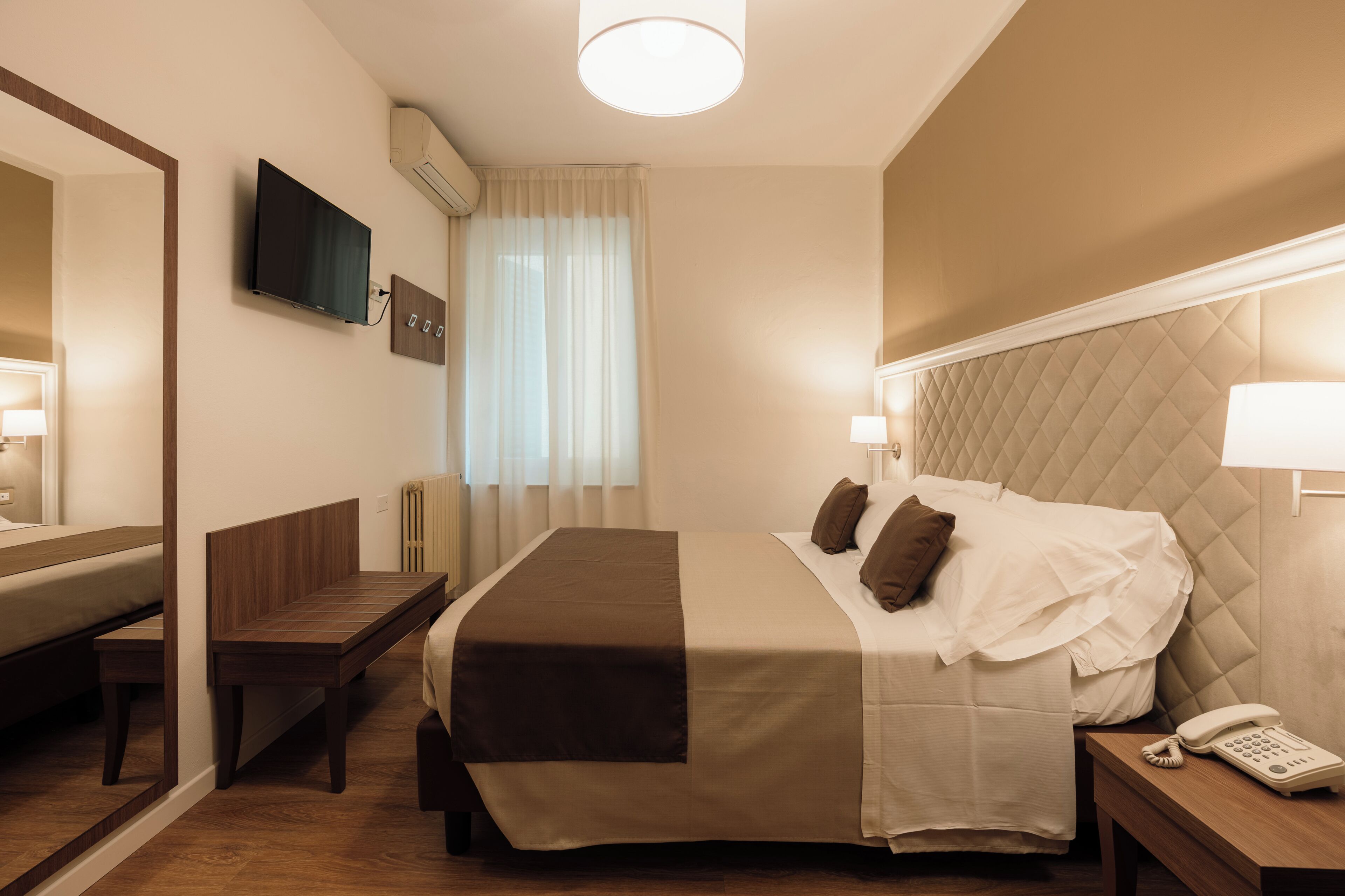 Een hotelkamer met een bed, nachtkastje, lamp, telefoon en een aan de muur bevestigde televisie.
